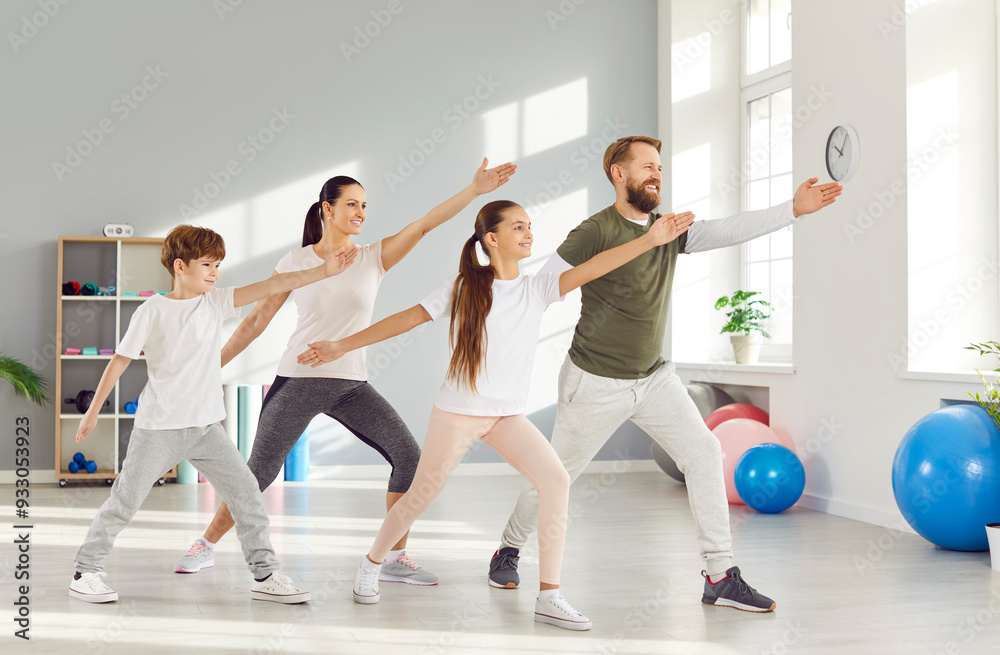 Parenthood and sport lifestyle. Happy active husband, wife and their two children doing family yoga session together. Family in comfortable clothes performs standing in same pose in bright home gym. Parenthood and sport lifestyle. Happy active husband, wife and their two children doing family yoga session together. Family in comfortable clothes performs standing in same pose in bright home gym.