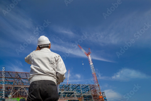 青空を背景にビル工事現場を見つめる作業着を着た男性技師の後ろ姿。 photo