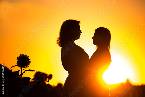 Silhouette of hugging mother and daughter in the rays of the sun against the backdrop of the sunset in a field of sunflowers. Dark photo