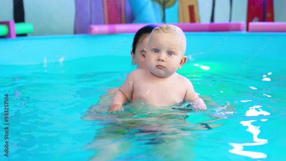Beautiful blond toddler is in the swimming pool. Female coach is holding is lifting a child up. Beautiful blond toddler is in the swimming pool. Female coach is holding is lifting a child up.
