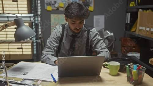 Focused man working on laptop in cluttered detective office with mug, files, and stationery photo