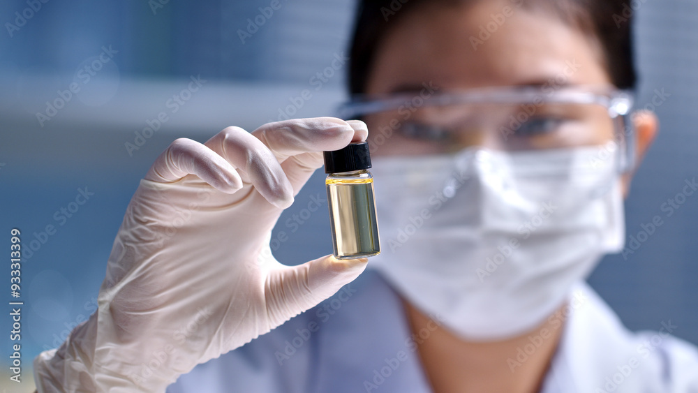 A light yellow vial featured in the center of photo frame by female scientist, over blurred laboratory scene. Front view, empty space for advertising and designing A light yellow vial featured in the center of photo frame by female scientist, over blurred laboratory scene. Front view, empty space for advertising and designing