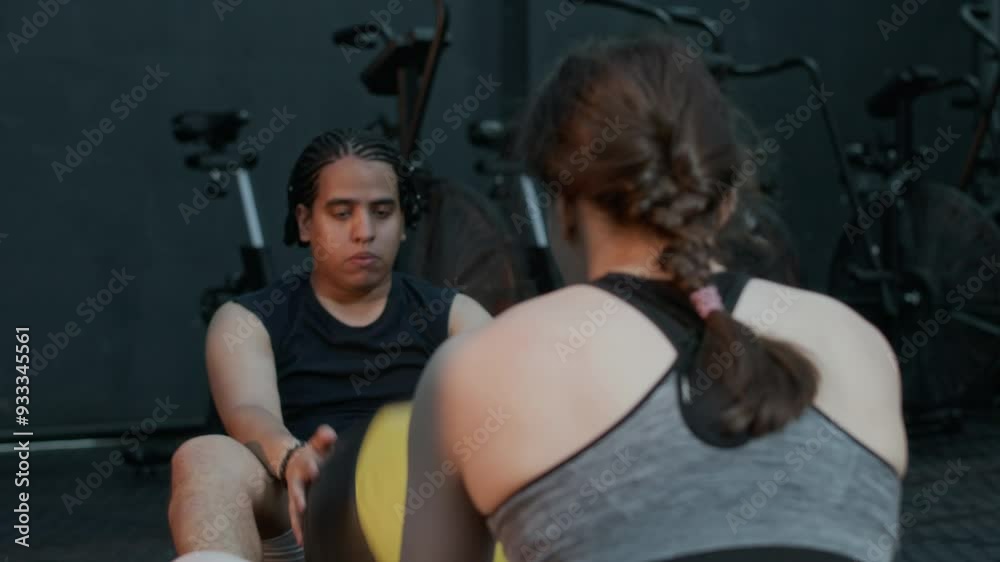 Tired young man passing med ball to his female instructor while sitting on floor and pumping abs during gym training Tired young man passing med ball to his female instructor while sitting on floor and pumping abs during gym training
