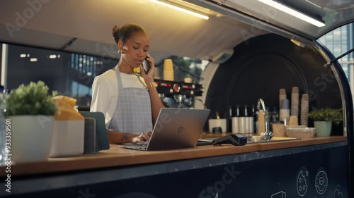 African American Female Food Truck Owner Multitasks, Using a Laptop Computer and Talking on a Phone. Small Business Manager Working Inside a Food Stand with Cups, Plants and Coffee Machines photo