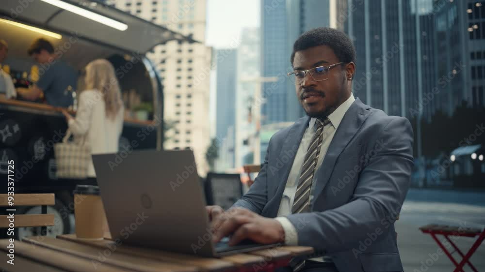 African Man Working on a Laptop Computer at a Street Cafe, Enjoying a Cup of Coffee. City Life with a Modern Food and Drinks Stand with Diverse People Ordering Lunch in the Background African Man Working on a Laptop Computer at a Street Cafe, Enjoying a Cup of Coffee. City Life with a Modern Food and Drinks Stand with Diverse People Ordering Lunch in the Background