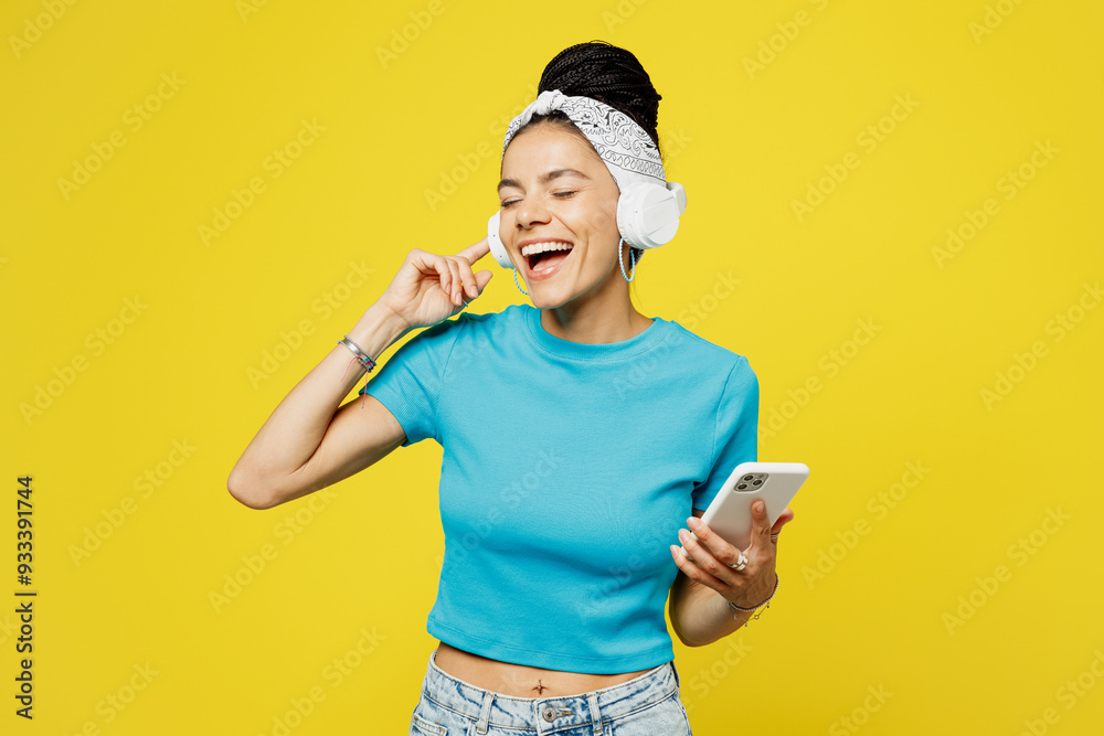 Young Latin woman she wears blue top t-shirt headband casual clothes listen to music in headphones use mobile cell phone sing song in microphone isolated on plain yellow background. Lifestyle concept. Young Latin woman she wears blue top t-shirt headband casual clothes listen to music in headphones use mobile cell phone sing song in microphone isolated on plain yellow background. Lifestyle concept.