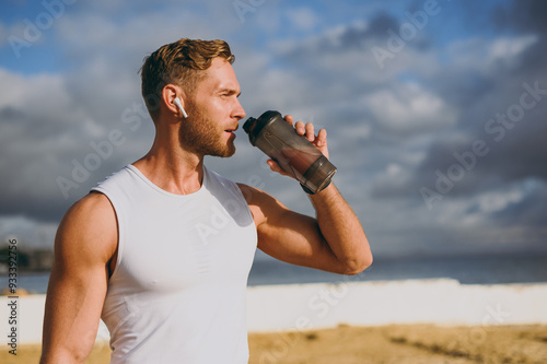 Foto Young cool strong athletic toned fit sportsman man wears sports clothes earphones drink protein shake warm up train at sunrise sun dawn over sea beach outdoor on pier seaside in summer day morning