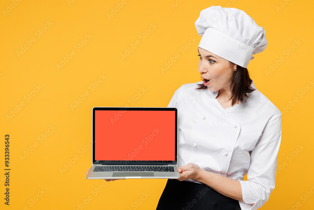 Young fun chef cook baker woman wear white shirt black apron uniform toque chefs hat hold use blank screen laptop pc computer look for recipe isolated on plain yellow background. Cooking food concept. Young fun chef cook baker woman wear white shirt black apron uniform toque chefs hat hold use blank screen laptop pc computer look for recipe isolated on plain yellow background. Cooking food concept.