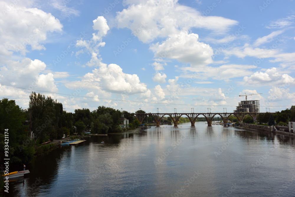 Beautiful view of the wide and long blue Dnipro River. Landscapes, a gray brick embankment with green trees, new construction sites and a beautiful bridge in the city of Dnipro. Beautiful view of the wide and long blue Dnipro River. Landscapes, a gray brick embankment with green trees, new construction sites and a beautiful bridge in the city of Dnipro.