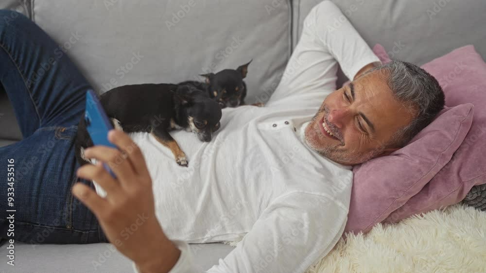 Middle-aged man relaxing on a sofa with two chihuahuas in a cozy living room setting, using a smartphone. Middle-aged man relaxing on a sofa with two chihuahuas in a cozy living room setting, using a smartphone.