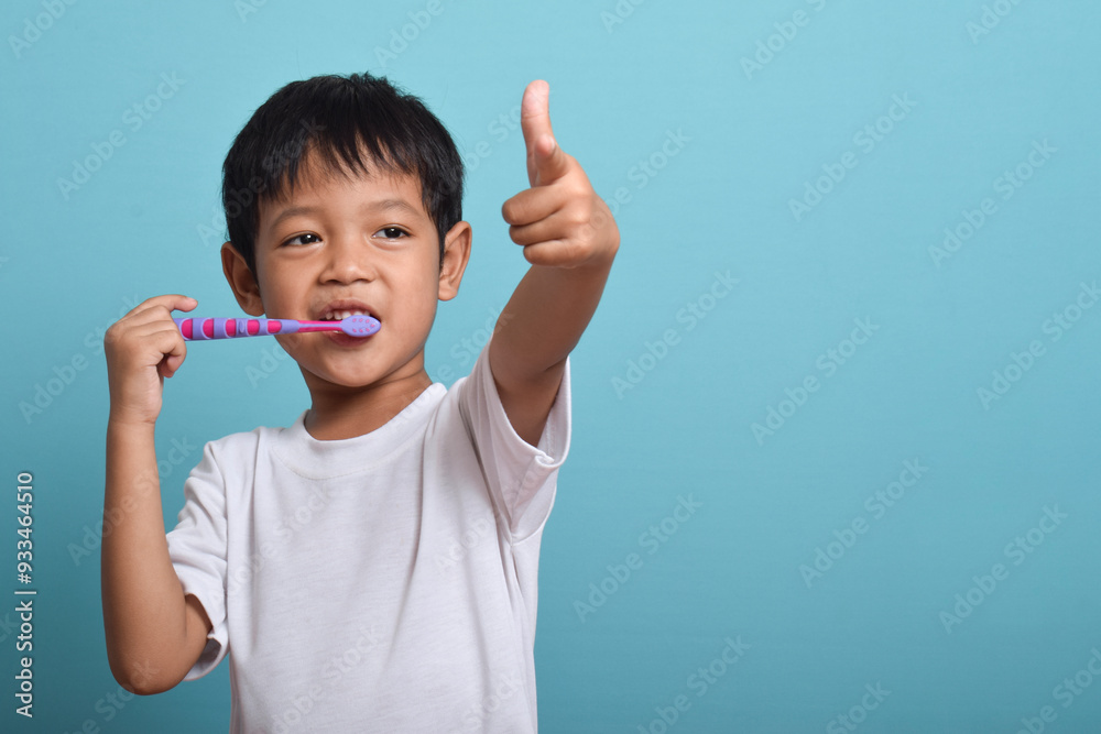 A happy Asian boy with clean teeth is brushing his teeth while pointing at the camera. Isolated on blue background. Health care, oral hygiene A happy Asian boy with clean teeth is brushing his teeth while pointing at the camera. Isolated on blue background. Health care, oral hygiene