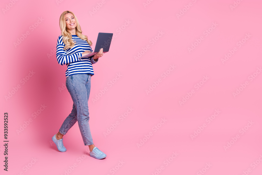 Full body photo of attractive mature walking hold netbook dressed stylish striped clothes isolated on pink color background Full body photo of attractive mature walking hold netbook dressed stylish striped clothes isolated on pink color background