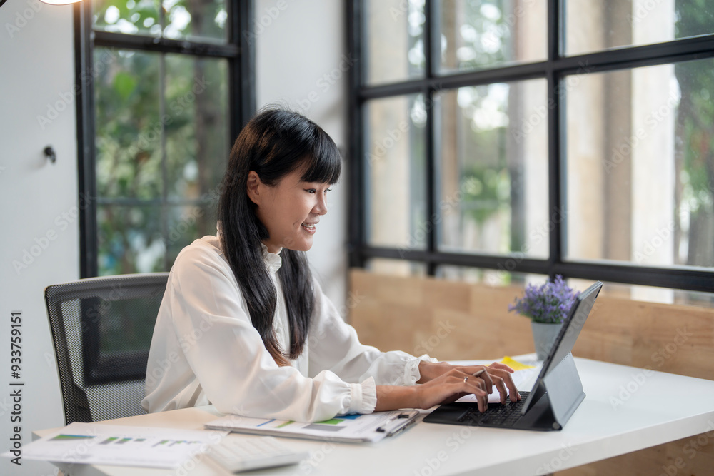 Young asian businesswoman smiling and typing on a tablet in a modern office Young asian businesswoman smiling and typing on a tablet in a modern office