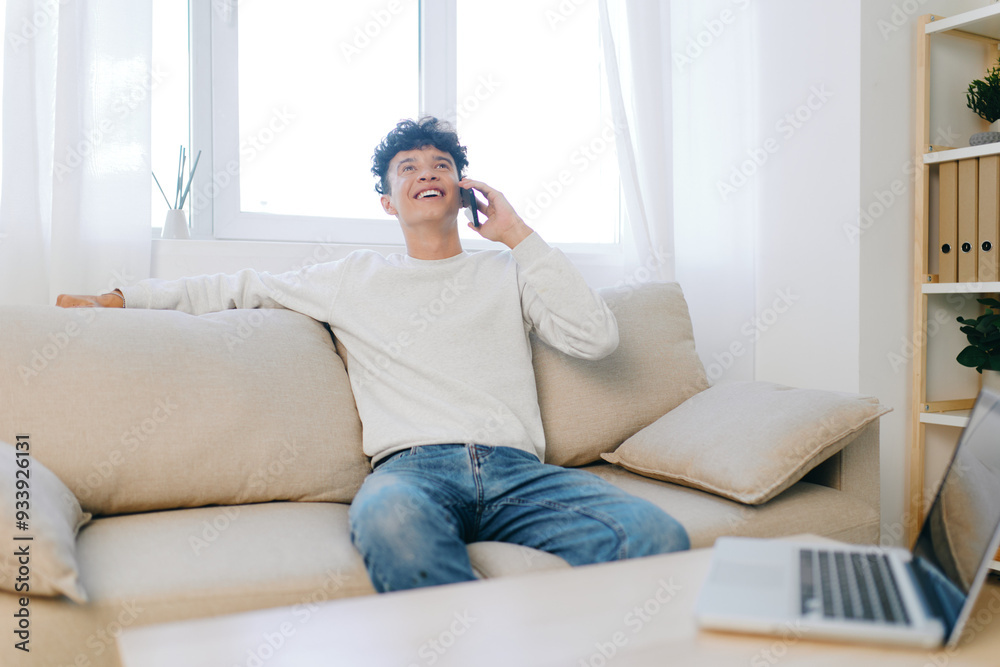 Young man talking on the phone while sitting on a comfortable couch in a bright, modern living room with a natural light background Relaxed and cheerful atmosphere, cozy design Young man talking on the phone while sitting on a comfortable couch in a bright, modern living room with a natural light background Relaxed and cheerful atmosphere, cozy design