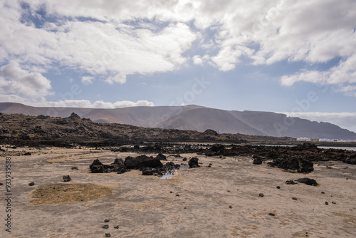 White sand beach with black rocks of volcanic origin. Mountains in the background and sky with big white clouds. Caletón Blanco, Lanzarote, Canary Islands, Spain