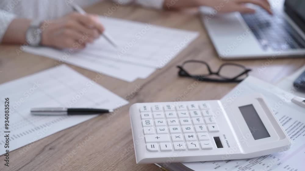 Close-up of a a calculator and documents on a wooden desk. Business professional working with papers and laptop on the background Close-up of a a calculator and documents on a wooden desk. Business professional working with papers and laptop on the background
