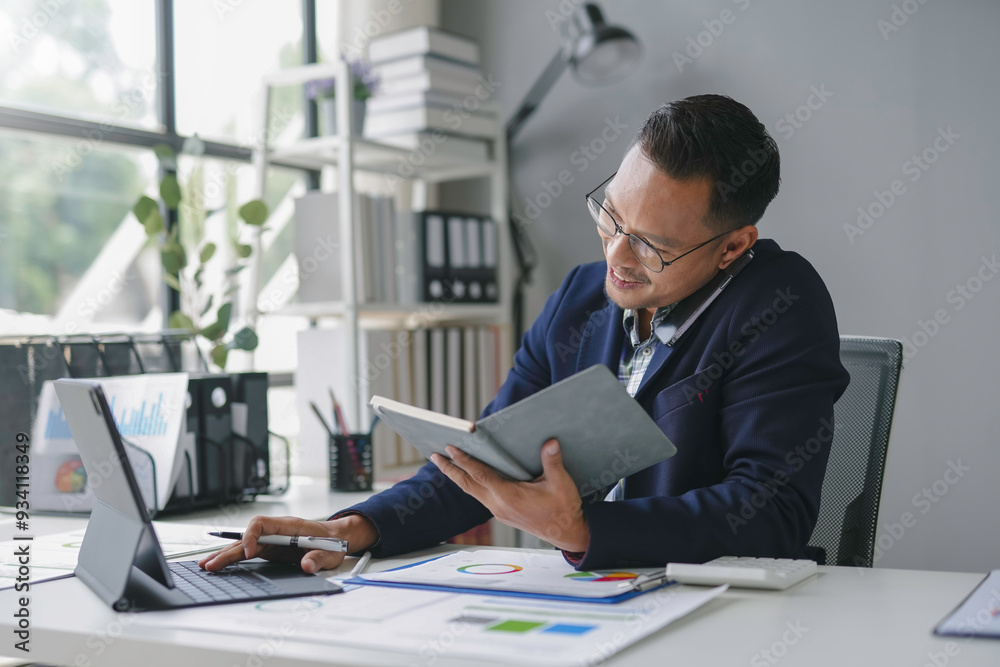 Fototapeta premium Asian accountant is working in his office, checking some numbers on a digital tablet and reading a book