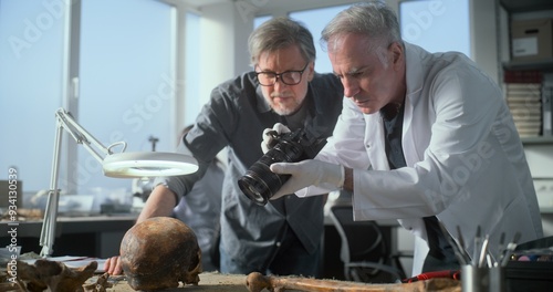 Archaeological laboratory: Senior paleoanthropologist and mature archaeologist take picture of fossil remains of prehistoric man skull with professional camera. Female scientist work in background. photo