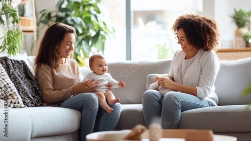 Two women interact joyfully with a baby on a cozy sofa in a sunlit living room filled with plants and natural light
