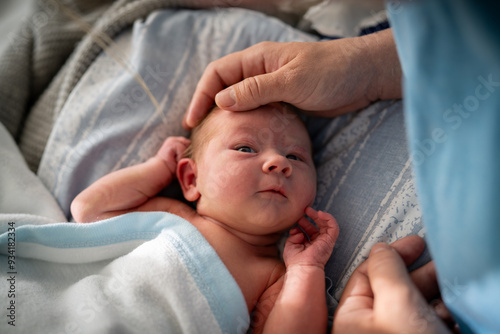 An infant wrapped in a blue blanket, lying down and looking upwards at their mother. The mothers hand gently touches the babys head, capturing a moment of care and bonding.  photo