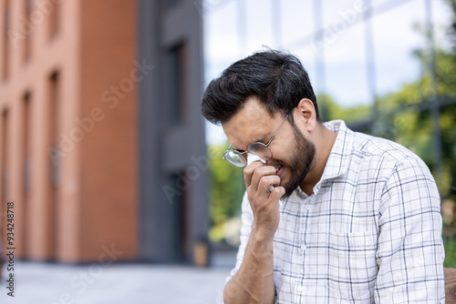 Close-up photo of a young sick Indian man sitting outside on the street near an office center and wiping his nose with a tissue, sneezing and covering his mouth with his hand