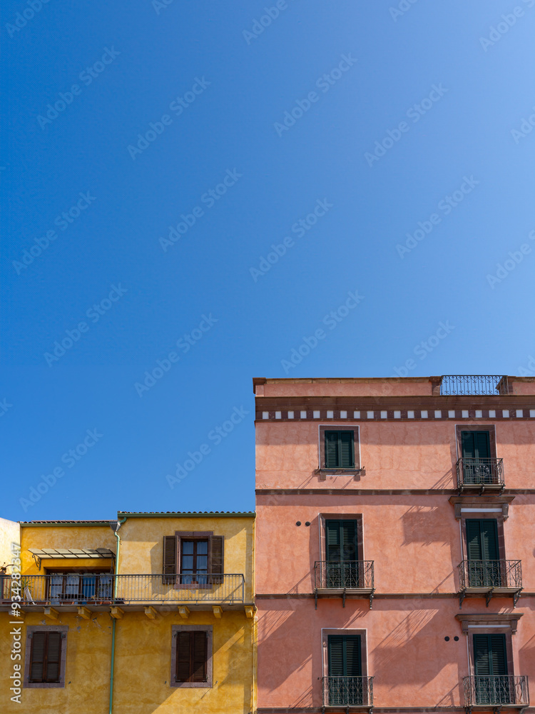 The facades of two adjacent buildings, one red and one yellow, create a perfect color triad against the blue sky. Located in Bosa, Italy, with Italian-style windows and Venetian shutters The facades of two adjacent buildings, one red and one yellow, create a perfect color triad against the blue sky. Located in Bosa, Italy, with Italian-style windows and Venetian shutters
