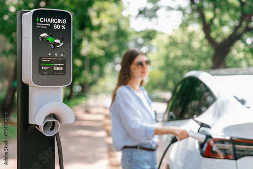 Young woman recharging battery for electric car during road trip travel EV car in natural forest or national park. Eco friendly travel during vacation and holiday. Exalt photo