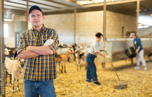 Confident positive male farmer, engaged in breeding dairy goats, posing in goat shed ..