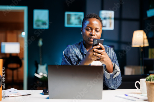 African american female blogger texting on mobile device while sitting in front of laptop on desk. Young black woman holding and using her smartphone, browsing the internet and social media.