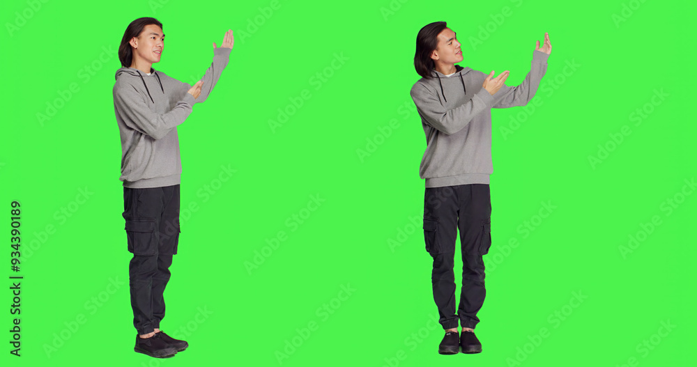 Asian man doing advertisement in studio, pointing at something next to him for new ad slogan. Young person smiling and working on promotional marketing, standing over full body greenscreen backdrop. Asian man doing advertisement in studio, pointing at something next to him for new ad slogan. Young person smiling and working on promotional marketing, standing over full body greenscreen backdrop.