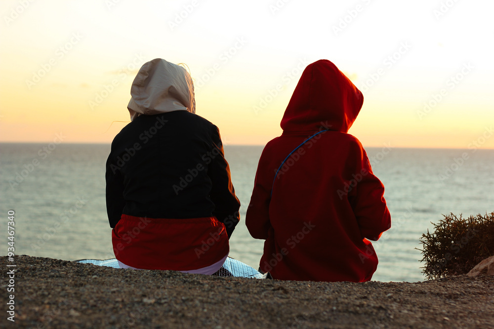 Close-up of two people sitting on a cliff overlooking the ocean at sunset. Sunset view, ocean horizon, outdoor relaxation, nature escape, calm moment, serene coastline, scenic landscape.
