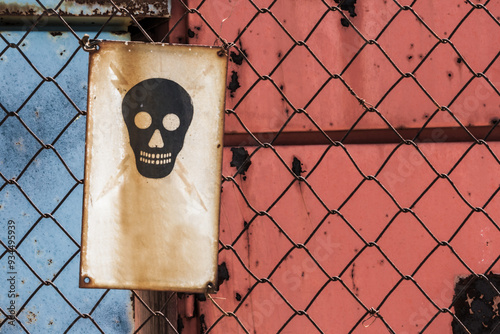 Obraz na plátně Weathered metal sign with a skull symbol warning of danger, attached to a rusty chain-link fence, creating an atmosphere of decay and caution in an industrial setting