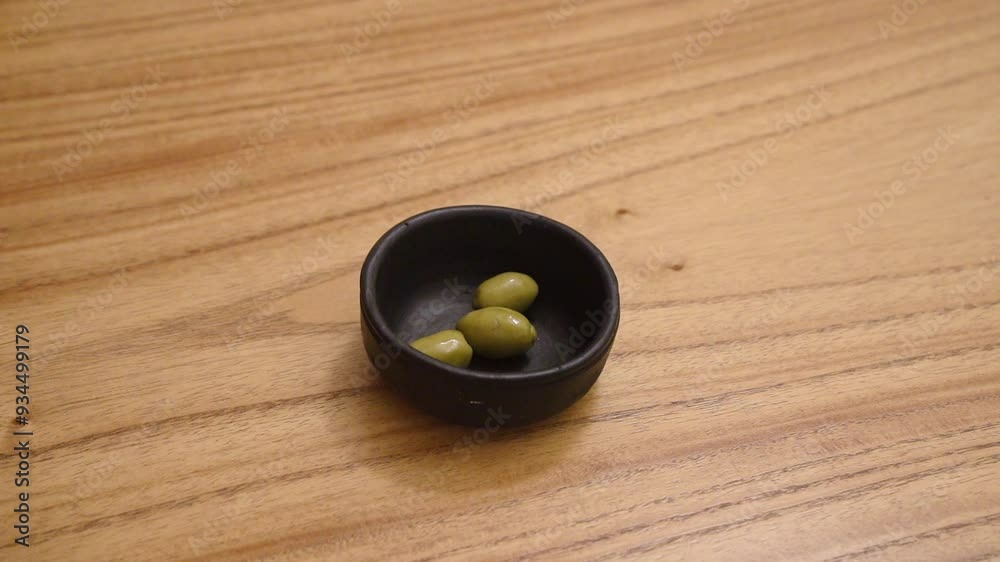 Closeup view of a man caucasian hand placing green olives in a black bowl on the wooden table Closeup view of a man caucasian hand placing green olives in a black bowl on the wooden table