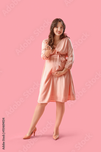 Young pregnant woman showing heart gesture on pink background. Valentine's Day celebration