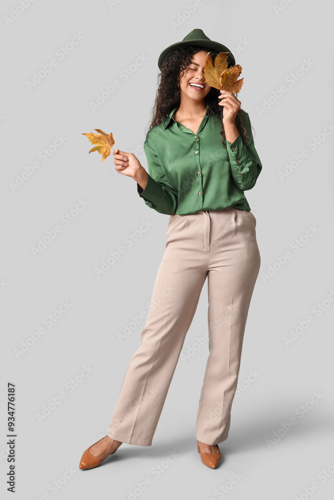 Beautiful young happy African-American woman in stylish outfit with autumn leaves on grey background Beautiful young happy African-American woman in stylish outfit with autumn leaves on grey background