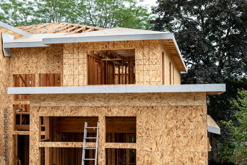 New large residential home construction in framing stage, with exterior walls covered in OSB sheathing, roof sheathing installation started over wood rafters, cloudy summer day
