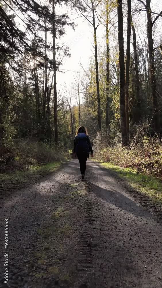 A woman with a backpack hikes down a gravel trail through a lush green forest. The sun is shining through the trees, creating a dappled lighting effect. British Columbia, Canada. A woman with a backpack hikes down a gravel trail through a lush green forest. The sun is shining through the trees, creating a dappled lighting effect. British Columbia, Canada.