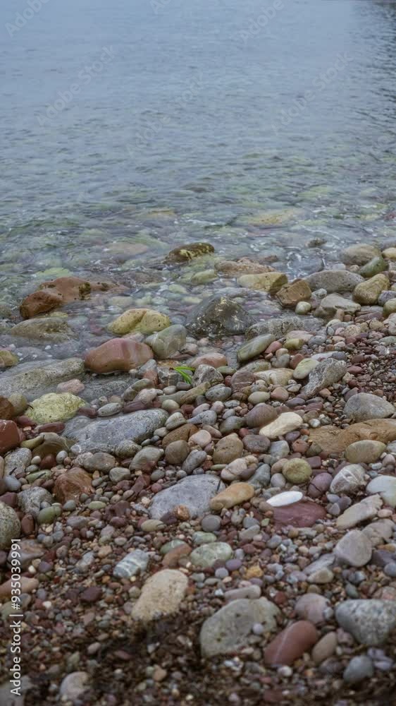 Pebbles along a rocky beach shoreline meeting the calm beach water on a cloudy day Pebbles along a rocky beach shoreline meeting the calm beach water on a cloudy day