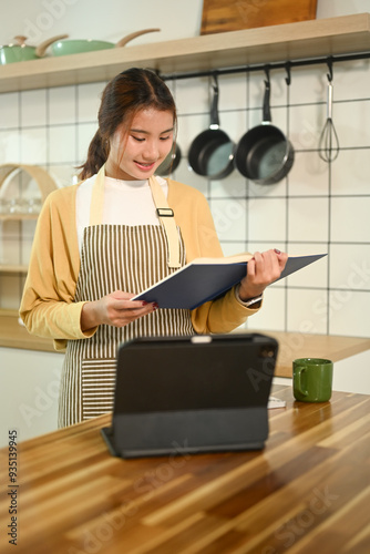 Beautiful young Asian woman wearing apron reading recipe book while preparing meal in kitchen