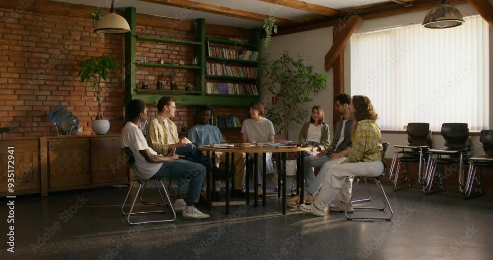 A group of young people are smiling and talking while sitting around a round table indoors. Notebooks and books on the table. Daylight A group of young people are smiling and talking while sitting around a round table indoors. Notebooks and books on the table. Daylight