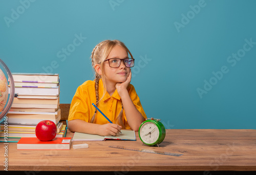 Back to school, cute girl sitting at a desk, doing her homework, witting, smiling. Blue background. 