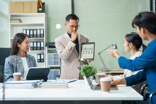 In a boardroom, an Asian team meets at a desk, presenting financial terms and strategies. Executives and employees collaborate, sharing ideas and work guidelines for success during an annual meeting.