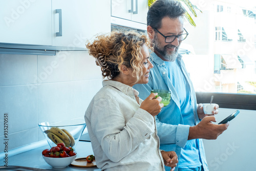 Adorable adult couple enjoying a morning breakfast together before starting their workday, sipping coffee and sharing smiles while looking at the same smartphone. A perfect depiction of a joyful, lovi photo