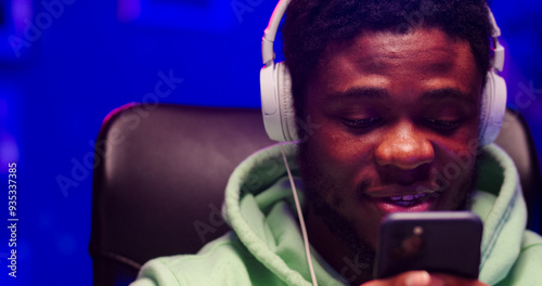 Close up portrait of satisfied African male wearing headphones holds phone in hands. Bearded face man listens to music during chatting with friend. photo