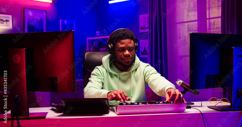 Close up portrait of young African male wearing headphones concentrates on creating music for party. Handsome bearded face man sits at table with different DJ equipment in LED lighted room. Close up portrait of young African male wearing headphones concentrates on creating music for party. Handsome bearded face man sits at table with different DJ equipment in LED lighted room.