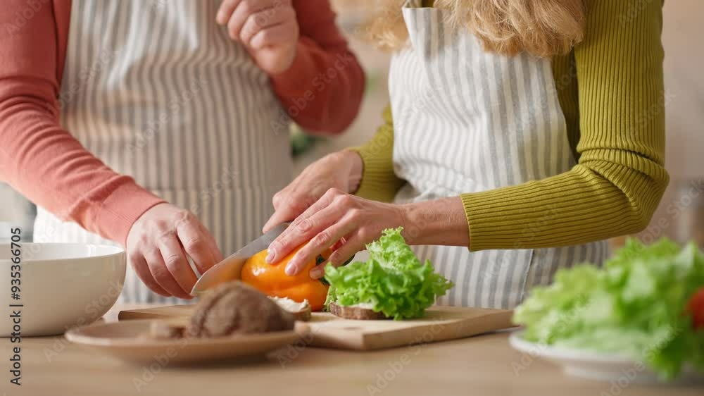 Woman with knife cutting fresh vegetables on cutting boards. Man helping his wife preparing sandwich in kitchen. People in aprons spending time together while cooking. Food preparation. Woman with knife cutting fresh vegetables on cutting boards. Man helping his wife preparing sandwich in kitchen. People in aprons spending time together while cooking. Food preparation.