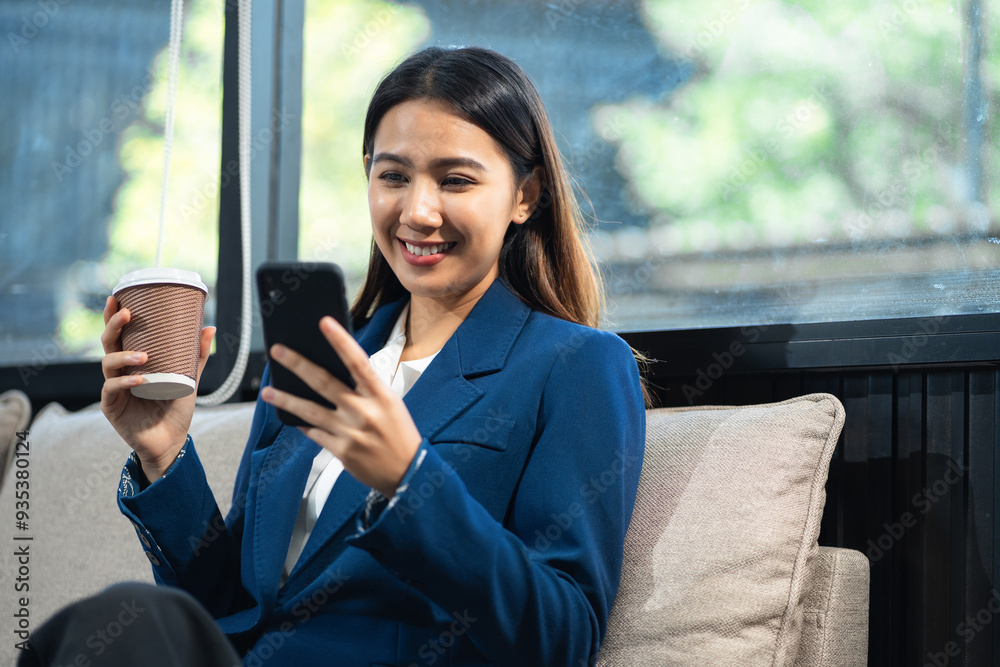 Obraz premium Asian businesswoman sits on sofa in a office working on laptop and enjoys a coffee.