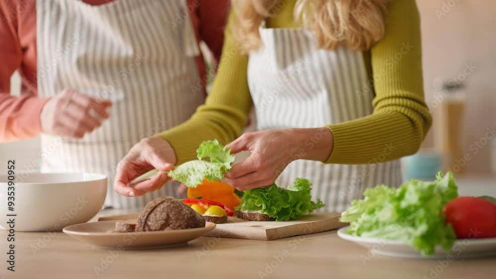 Camera view of man helping his wife cooking in kitchen. People wearing aprons while preparing food. Woman putting salad and vegetables on bread. Healthy nutrition. Making sandwiches or appetizers. Camera view of man helping his wife cooking in kitchen. People wearing aprons while preparing food. Woman putting salad and vegetables on bread. Healthy nutrition. Making sandwiches or appetizers.