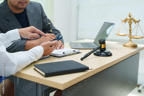 Fototapeta Naklejka Na Ścianę i Meble -  Thai lawyer in a suit works on a laptop at his office desk, with a gavel beside him. intersection of technology and law, representing professionalism, legal expertise, and corporate business.