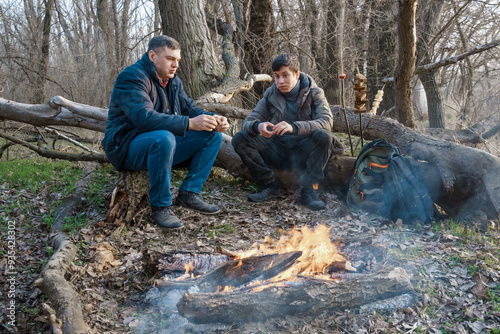 Two men make a campfire for cooking in the forest, father and son sitting on a log while hiking and outdoor activities, early spring landscape photo